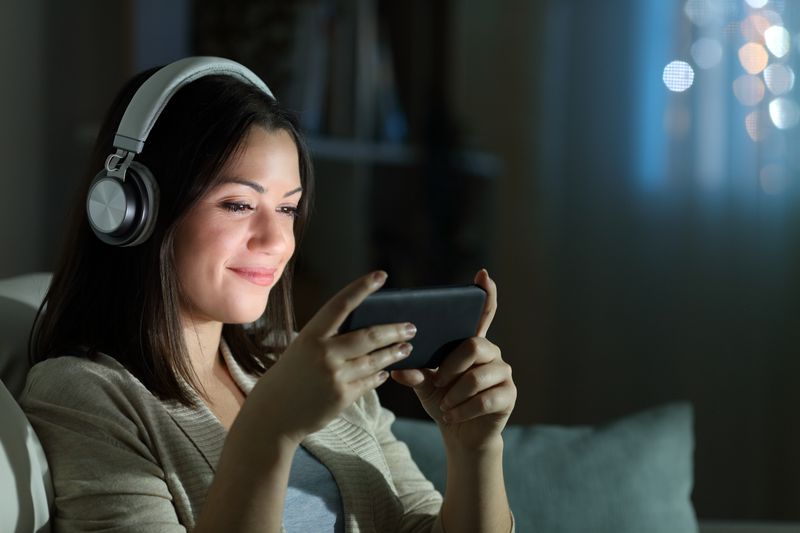 Relaxed woman with headphones watching video on smart phone in the night sitting on a couch in the living room at home