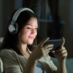 Relaxed woman with headphones watching video on smart phone in the night sitting on a couch in the living room at home