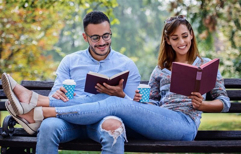 An overly happy couple sit and recline on a park bench while reading. The couple are intentionally or otherwise wearing the same colour denim jeans and shirts, while holding identical blue and white spotted coffee cups and are reading burgundy coloured books. 