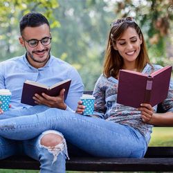 An overly happy couple sit and recline on a park bench while reading. The couple are intentionally or otherwise wearing the same colour denim jeans and shirts, while holding identical blue and white spotted coffee cups and are reading burgundy coloured books. 