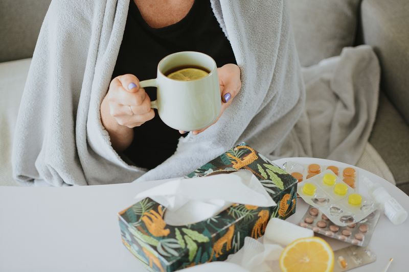 woman drinks tea with a lemon slice while wrapped in a blanket (only her torso is visible); on the table in front of her is a box of tissues, a selection of lozenges and pills, a nasal spray and the rest of the lemon