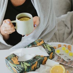 woman drinks tea with a lemon slice while wrapped in a blanket (only her torso is visible); on the table in front of her is a box of tissues, a selection of lozenges and pills, a nasal spray and the rest of the lemon