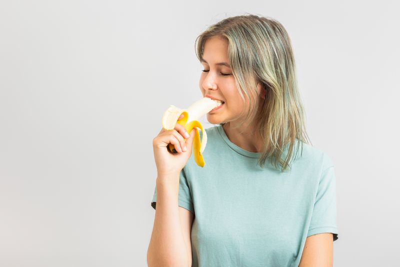 woman on beige background wearing mint green t-shirt with blonde hair biting into a banana