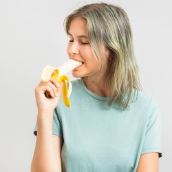 woman on beige background wearing mint green t-shirt with blonde hair biting into a banana