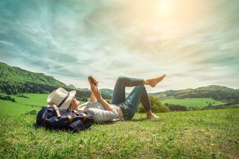 Woman holding a phone lounging on sunny grassy hill, next to a backpack