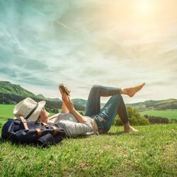 Woman holding a phone lounging on sunny grassy hill, next to a backpack