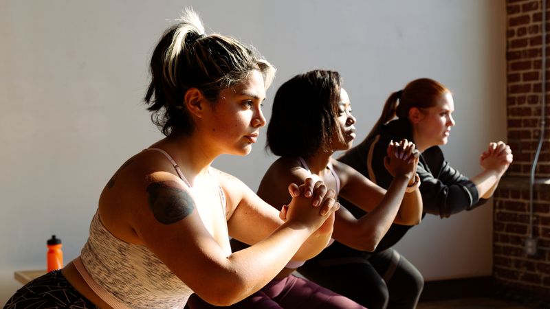 Three women working out in a gym. They are pictured from the waist up, standing in a line facing towards the right. They are of diverse ethnicities and body types and appear to be performing squats in an exercise studio, wearing workout clothes.