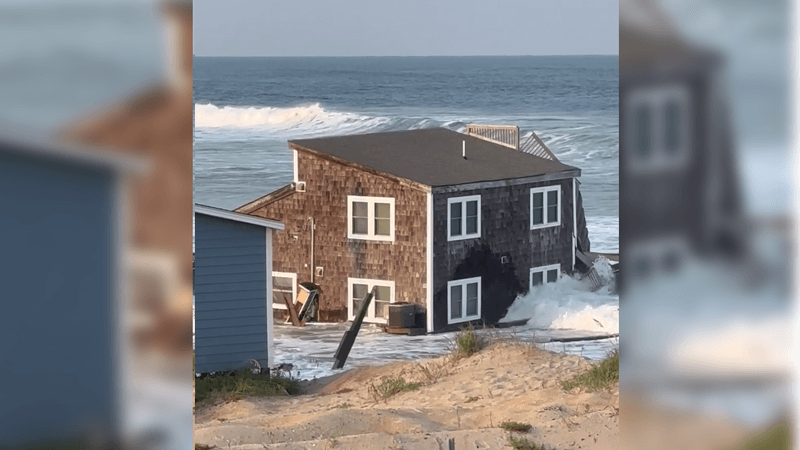 The remains of a collapesed beach house in the edge of the water at the ocean surrounded by waves. There are sand dunes in the foreground.