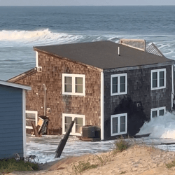 The remains of a collapesed beach house in the edge of the water at the ocean surrounded by waves. There are sand dunes in the foreground.