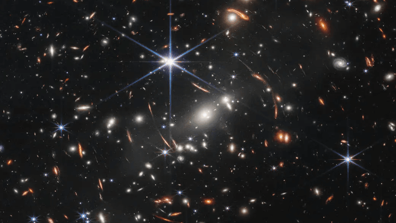 In the center of the image, between 4 o’clock and 6 o’clock in the bright star’s spikes, are several bright, white galaxies.  These are members of the galaxy cluster.  The background of space is black. Thousands of small galaxies appear across the image. Their colors vary. Some are shades of orange, others are white. Most appear as fuzzy ovals, but a few have distinctive spiral arms. There are also many thin, long, orange arcs. They follow invisible concentric circles that curve around the center of the image. These are images of background galaxies that have been stretched and distorted by the foreground galaxy cluster.