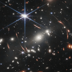 In the center of the image, between 4 o’clock and 6 o’clock in the bright star’s spikes, are several bright, white galaxies.  These are members of the galaxy cluster.  The background of space is black. Thousands of small galaxies appear across the image. Their colors vary. Some are shades of orange, others are white. Most appear as fuzzy ovals, but a few have distinctive spiral arms. There are also many thin, long, orange arcs. They follow invisible concentric circles that curve around the center of the image. These are images of background galaxies that have been stretched and distorted by the foreground galaxy cluster.