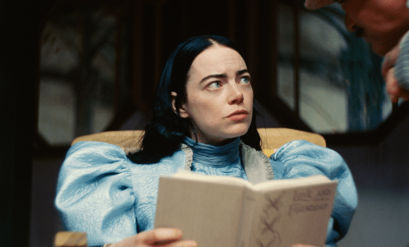 Emma Stone as Bella Baxter in Poor Things, wearing a blue puff-sleeved dress, reading a book, and glancing into the top right corner of the screen