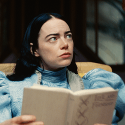 Emma Stone as Bella Baxter in Poor Things, wearing a blue puff-sleeved dress, reading a book, and glancing into the top right corner of the screen