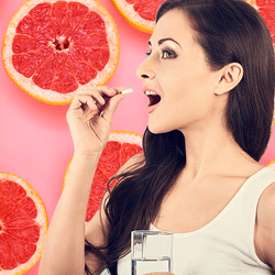 woman with long dark hair taking a pill holding a glass of water, against a backgroun of a repeating pattern of pink grapefruit slices against a light pink background