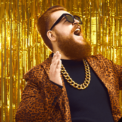 man with red hair and beard dancing against a wall of gold streamers, wearing sunglasses, a gold chain and a leopard print blazer; there is a diagonal black line dividing the image from top right to bottom left, and on the other side the same man lies in bed facing the camera, wearing white PJs and a sleeping mask and holding a teddy