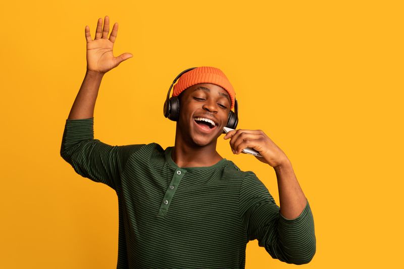 Man in orange beanie, black headphones, and a green jumper, singing with their eyes closed in front of a mustard yellow background