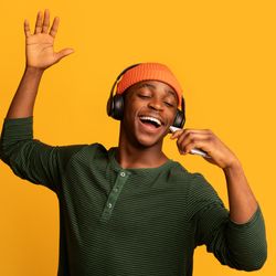 Man in orange beanie, black headphones, and a green jumper, singing with their eyes closed in front of a mustard yellow background