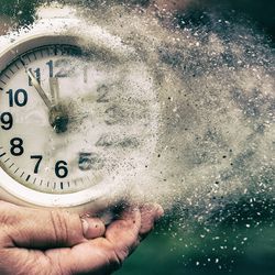 Person holding a white clock in both hands, the clock is dissolving into dust and blowing away to the right