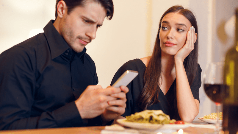 A man ignoring his date in favor of his smartphone. His date, a woman, looks annoyed and bored.