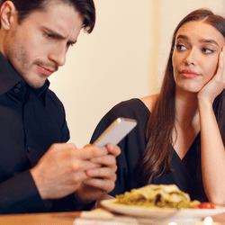 A man ignoring his date in favor of his smartphone. His date, a woman, looks annoyed and bored.