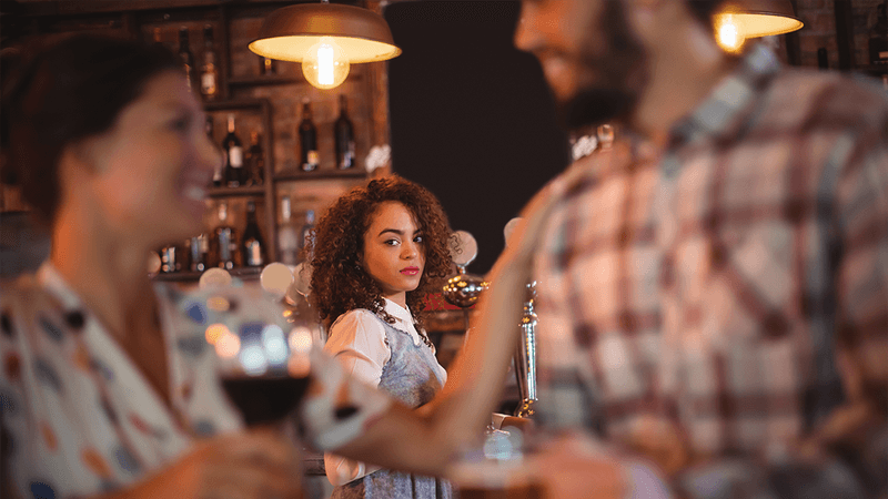 Two people flirting at a bar, a woman in the background looking jealous.