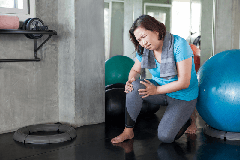 Woman kneeling in a gym clutching her knee in pain, wearing a blue T shirt and grey leggings with a towel around her shoulders